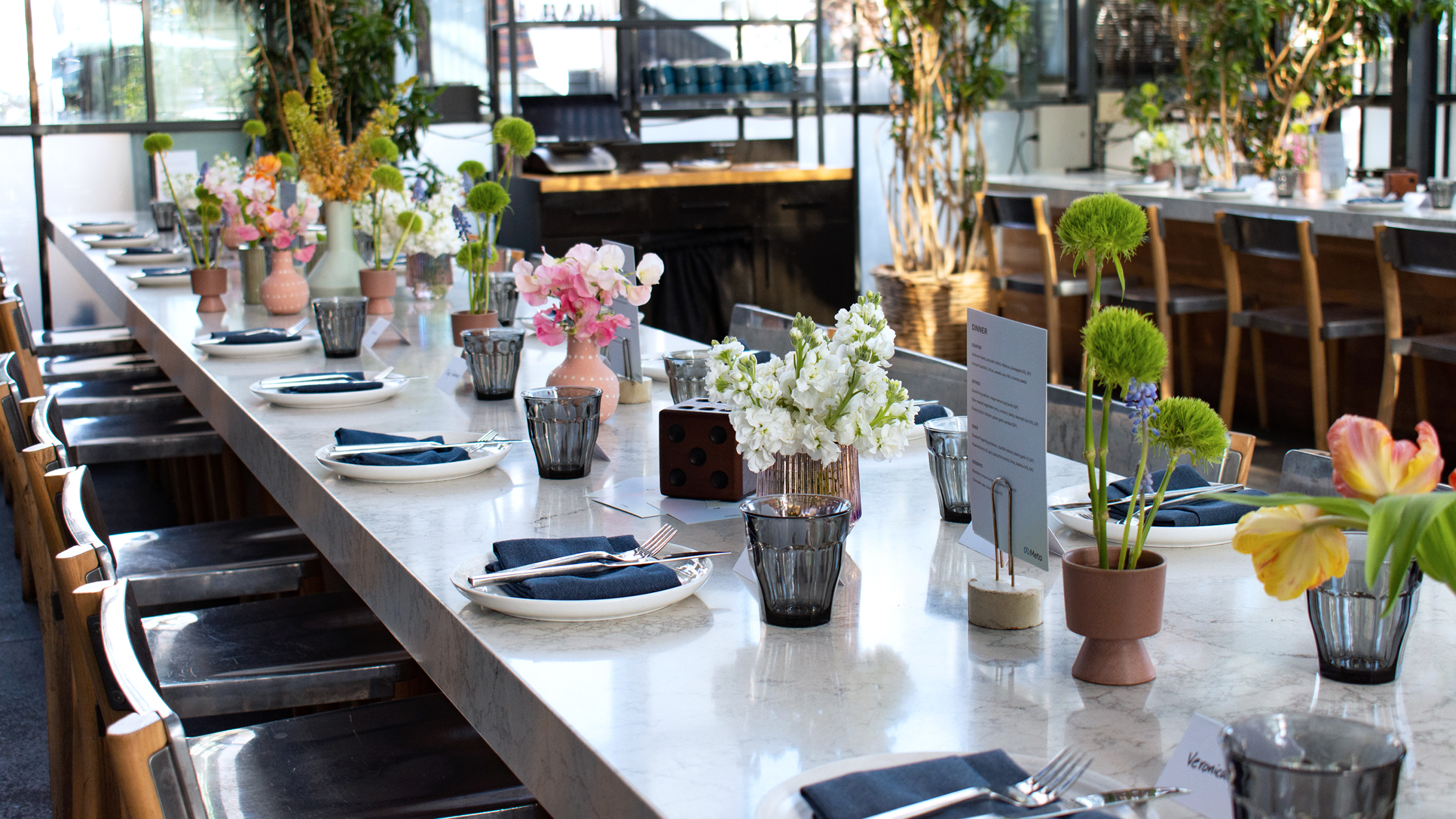 A long dining table set for a meal with a variety of colorful floral centerpieces and neatly arranged table settings including plates, napkins, and cutlery. Modern chairs line the table, and the restaurant interior features large windows and greenery.