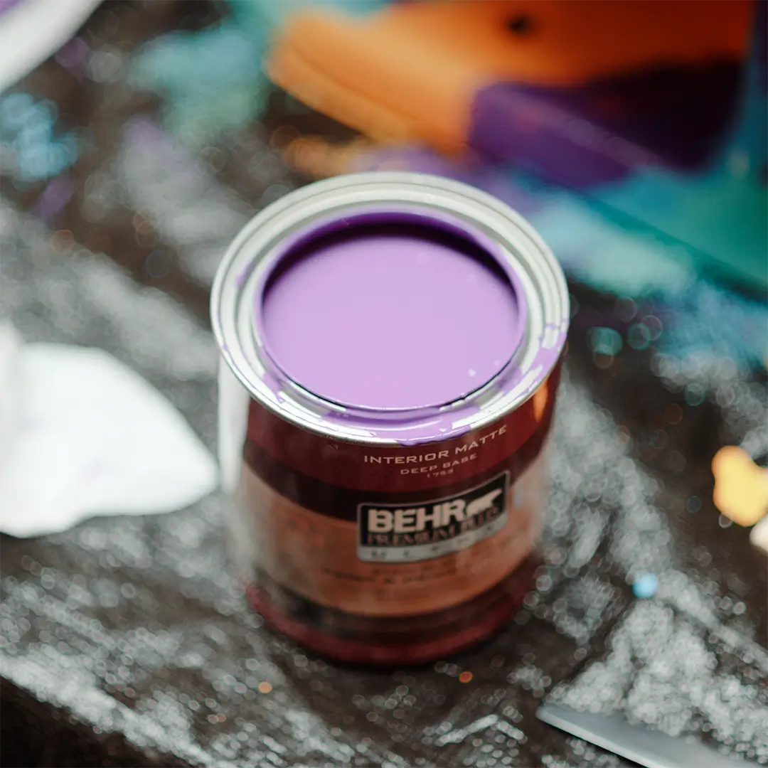 An open can of BEHR interior matte paint sits on a speckled, paint-splattered surface. The paint inside the can is a vibrant shade of purple. Partially visible tools and materials surround the can, suggesting an ongoing painting project.