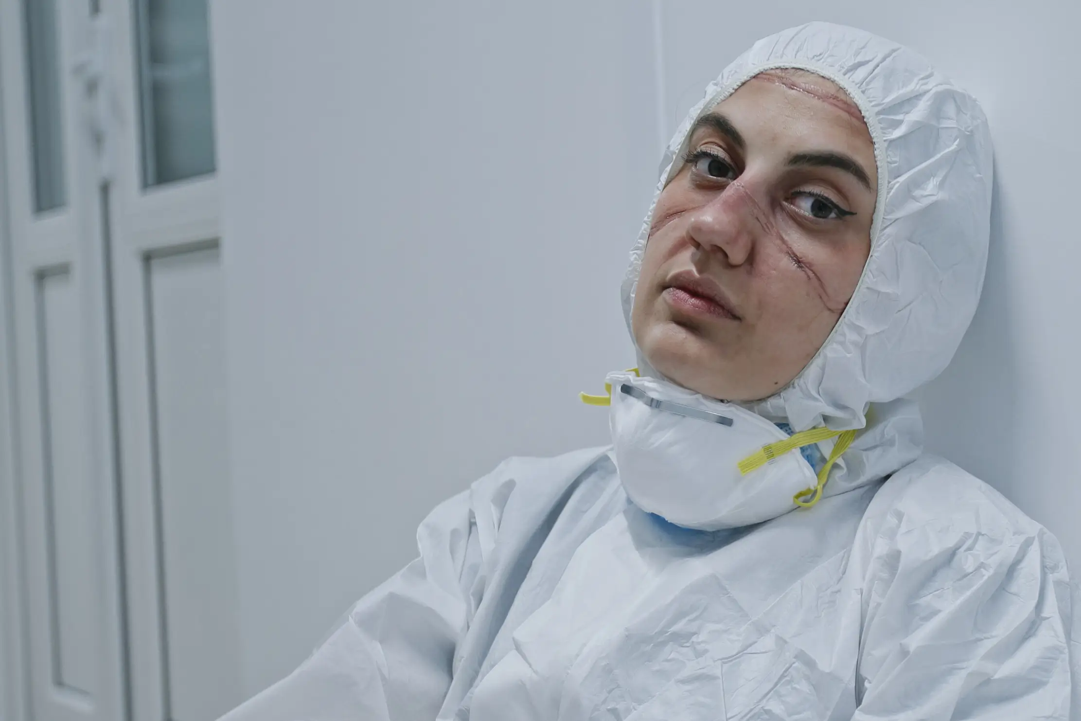 An exhausted healthcare worker wearing a white protective suit, face mask, and head covering sits against a wall, with noticeable facial marks from prolonged mask use. The setting appears to be a clinical or hospital environment, highlighting the urgent need for effective health care advocacy.