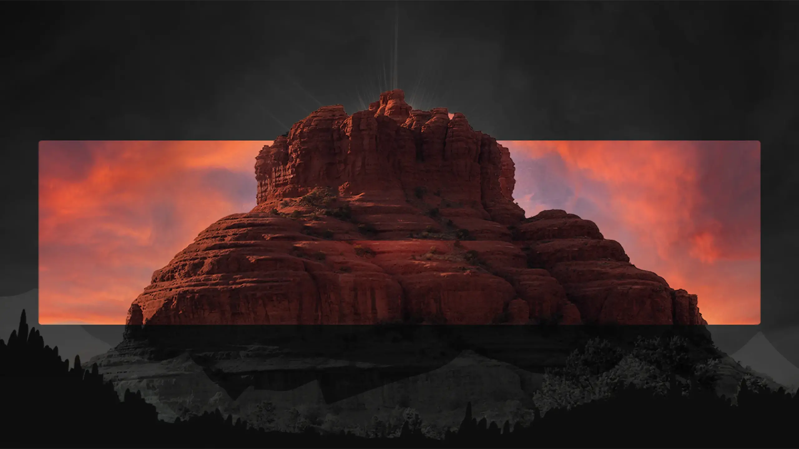 A dramatic landscape featuring a large red rock formation, illuminated by a setting or rising sun with a vivid, colorful sky in the background. The image is artistically framed with dark silhouettes of mountains in the foreground and a black border above and below, perfect for digital impressions.