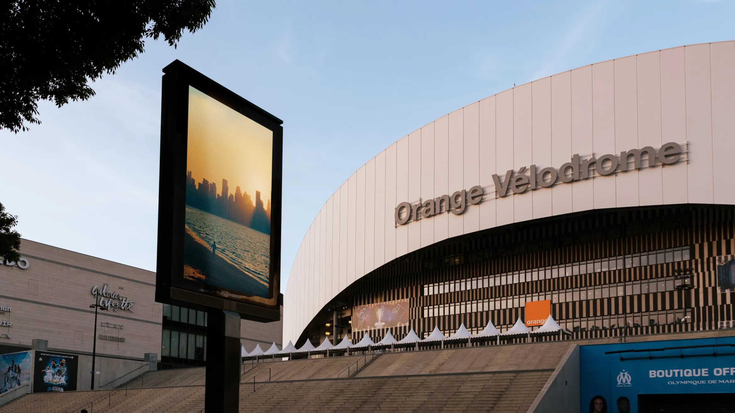 An outdoor scene shows the Orange Vélodrome stadium with its curved white facade and name prominently displayed. In the foreground to the left, there is a large vertical billboard featuring a serene image of a sunset over water with a city's skyline in the background.