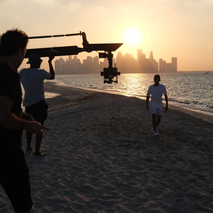 A man in white walks on a sandy beach at sunset towards the camera. Another man and a cameraman operate filming equipment in the foreground. The sun is setting behind the ocean, casting a golden glow and illuminating city buildings in the background.