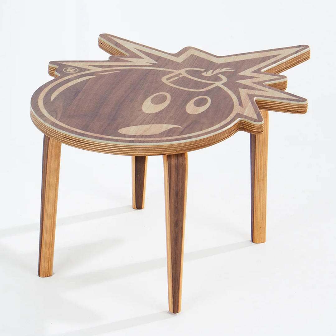 A small wooden table with three legs, featuring a unique tabletop design resembling a bold, cartoonish face with expressive eyes, a smiling mouth, and a spiky hair-like outline. The craftsmanship highlights the wood grain in varying shades.