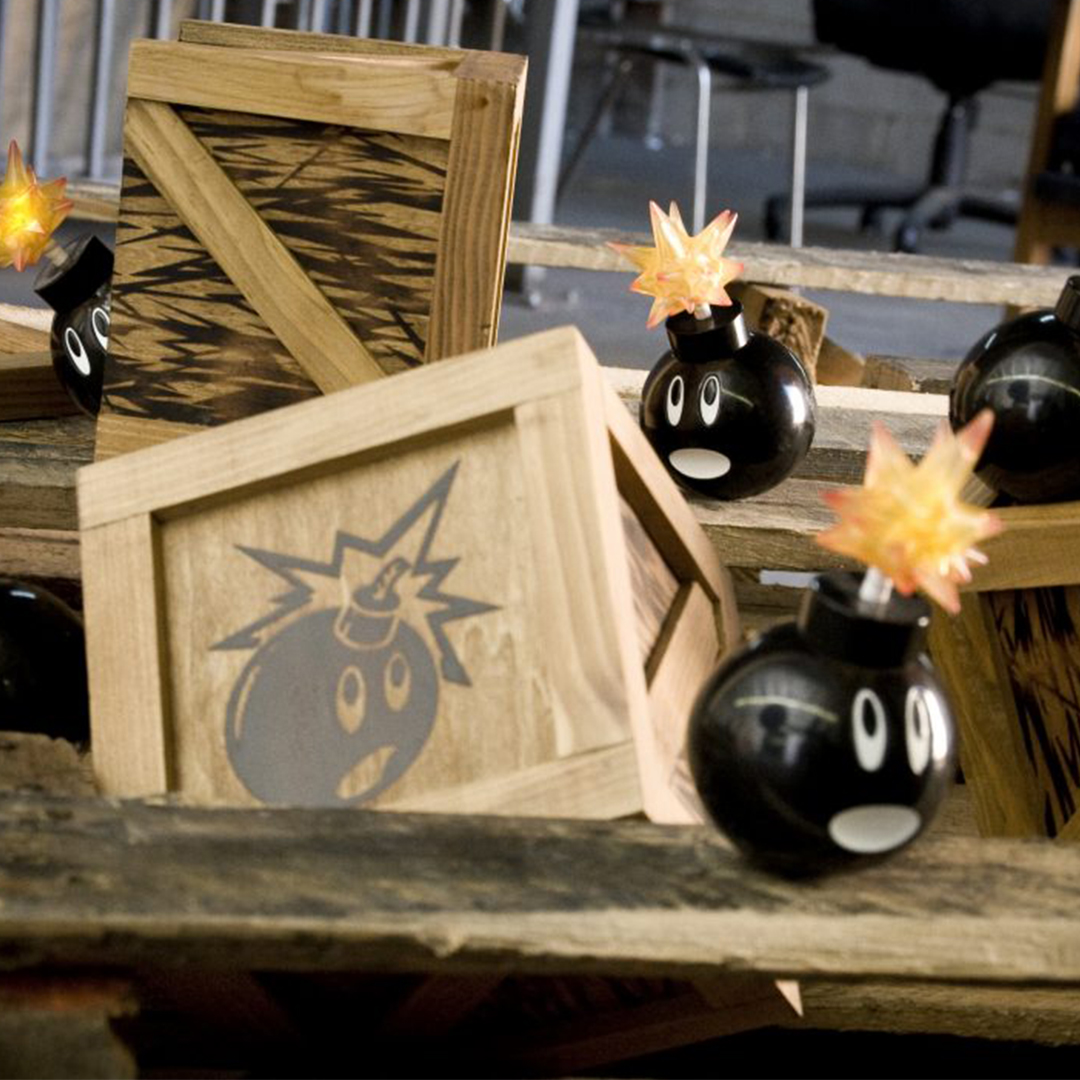 A group of black, cartoon-like bomb figures with expressive eyes and lit fuses are scattered among wooden crates. Some crates have a similar bomb face image printed on them. The setting appears to be a rustic indoor environment with wooden planks.