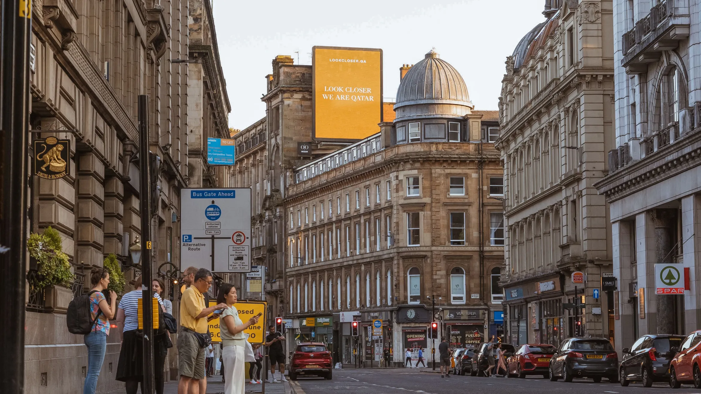 A bustling city street lined with historic buildings. Pedestrians walk along the sidewalks and gather on one side. Cars are parked along the street. An illuminated billboard at the end of the street reads, "LOOK CLOSER, WE ARE CAVTAR." Various street signs are visible.