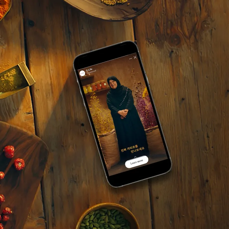 A smartphone rests on a rustic wooden table surrounded by spices and dried fruits. The phone screen shows a video of a person with a warm smile, wearing a dark outfit, standing inside a room with colorful decorations. The video includes a "Learn more" button.