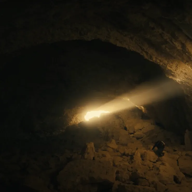 A lone explorer clad in dark clothing is seen navigating the rugged and rocky terrain of a dimly-lit cave with a small opening that casts a narrow beam of bright light onto the cave floor, highlighting the vast, shadowy interior of the cavern.