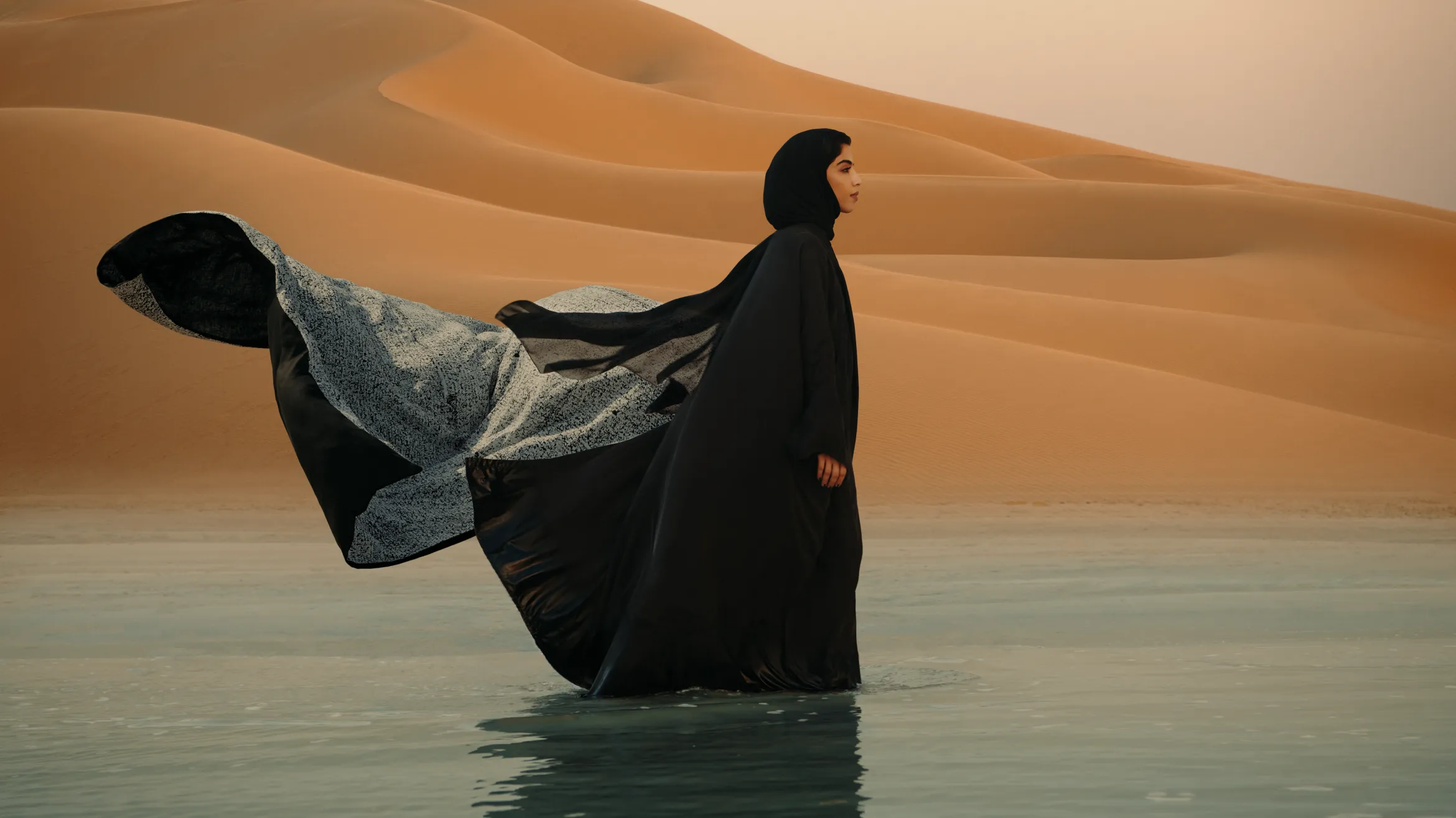 A person wearing a long, flowing black cloak walks through a shallow body of water in a desert landscape. The cloak billows behind them, contrasting with the golden sand dunes in the background against a soft, pastel sky.