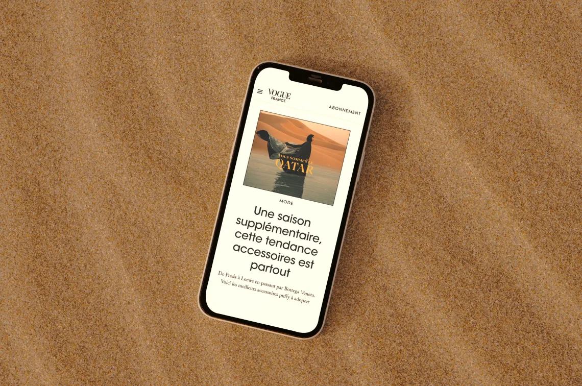 A smartphone displaying an article from Vogue magazine rests on a sandy surface. The screen shows an image of two women on a beach and text in French, with the headline "Une saison supplémentaire..." followed by more text about fashion trends and accessories.