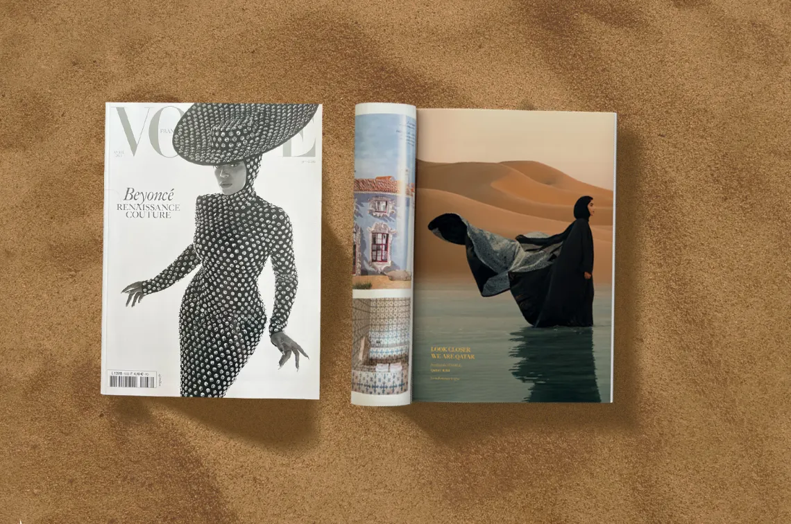 Two magazines lie open on sandy ground. The left magazine's cover features a black-and-white image of a person in a textured, form-fitting dress and a large, elaborate hat. The right magazine displays an open page with an image of a person wearing a flowing dark garment, walking through water in a desert landscape.