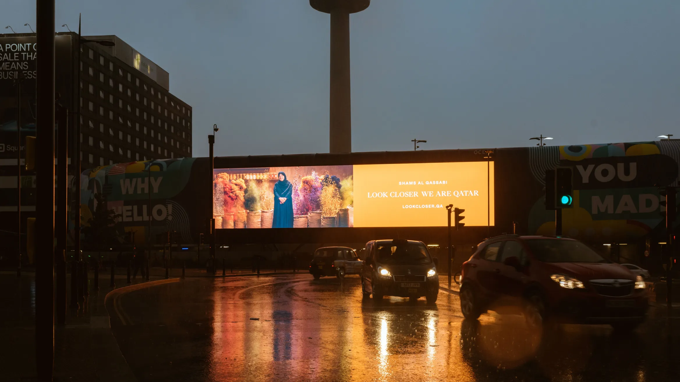 A city street at dusk with wet pavement reflecting lights. Cars drive past. A large digital billboard displays an advertisement that reads "LOOK CLOSER WE ARE QATAR" along with an image of a person standing in front of the Qatari flag. Buildings and a tower are in the background.