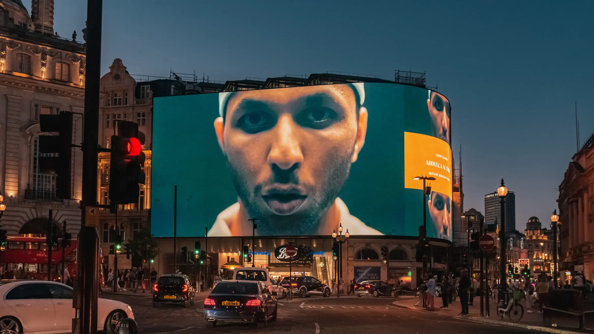 A bustling city street at dusk with cars and people. A large, illuminated digital billboard dominates the scene, displaying a close-up of a person's face with a somber expression. Historic buildings with lit windows surround the street, and the sky is a deep blue.