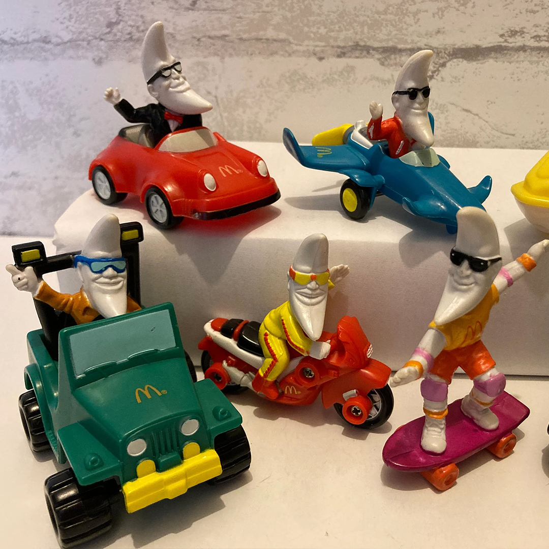 A collection of five toy figures featuring a character with a crescent moon-shaped head and sunglasses dressed in various outfits. They are riding a red car, a blue car, a green vehicle, a red motorcycle, and a purple skateboard.