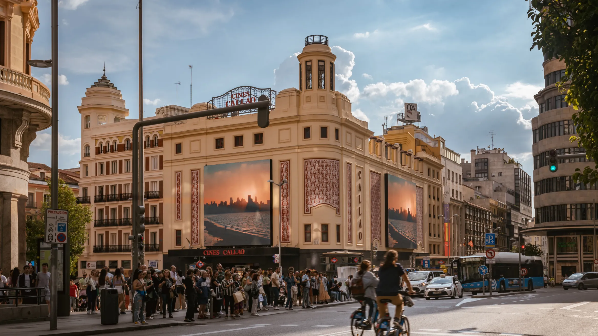 A bustling city scene shows a crowd of people gathered on a sidewalk in front of a grand, historic building adorned with billboards. Cyclists and pedestrians traverse the street, and vehicles, including buses, are visible. The sky is partly cloudy with soft sunlight.