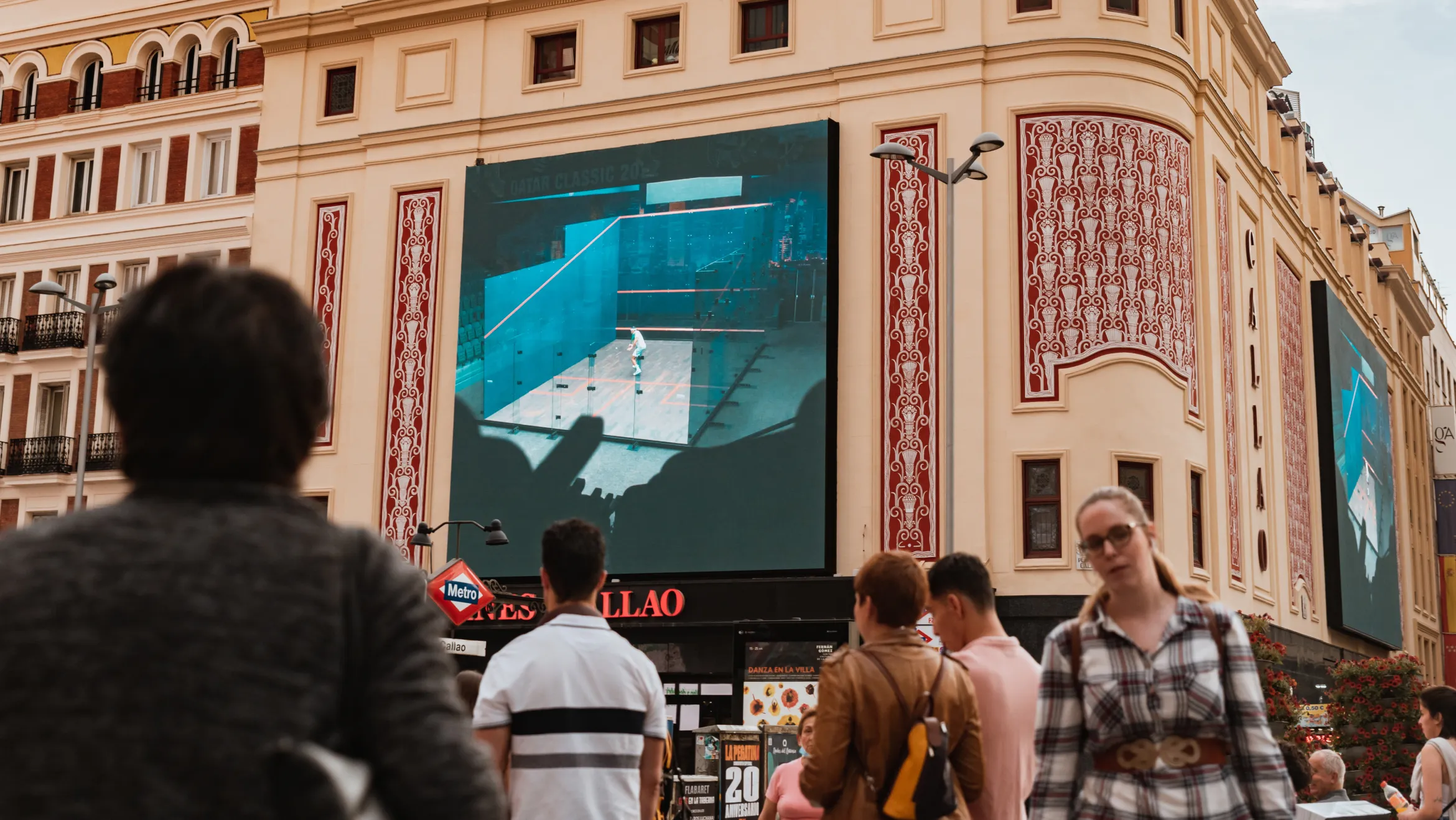 A large screen on the side of an ornate building displays an indoor pool with some surrounding architecture. Several people are gathered in the foreground, some facing away and some walking towards the camera. The scene takes place in a busy outdoor urban area.