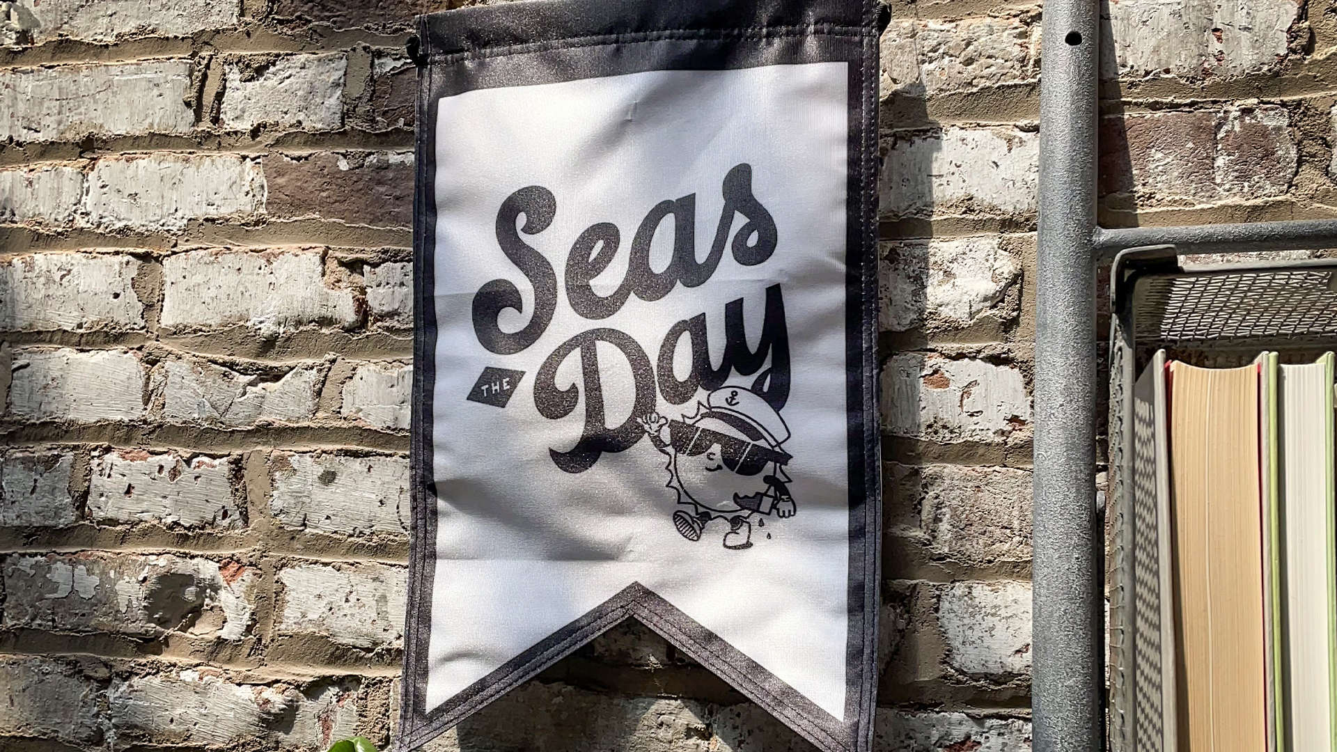 A white pennant with "Seas the Day" and a cartoon character wearing a sailor hat is hanging on a brick wall. The pennant is bordered in black, and there is a metal shelf holding books on the right side of the image.