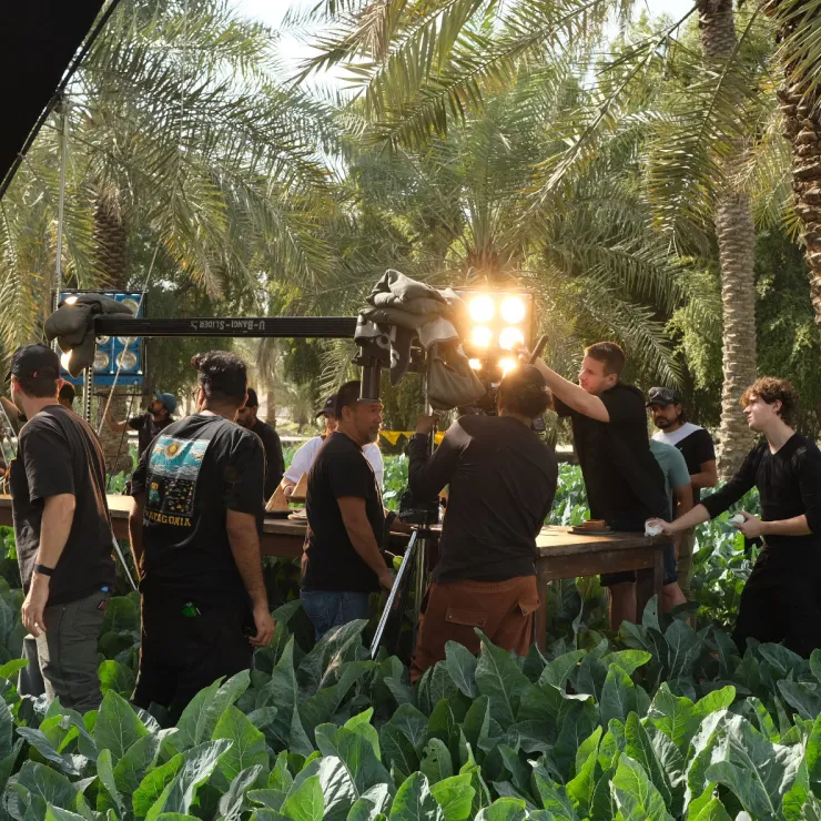 A group of people are gathered in a lush, green field with large leafy plants. They are involved in a filming setup, with one person operating a camera crane and others adjusting equipment and lighting. Tall palm trees are visible in the background.