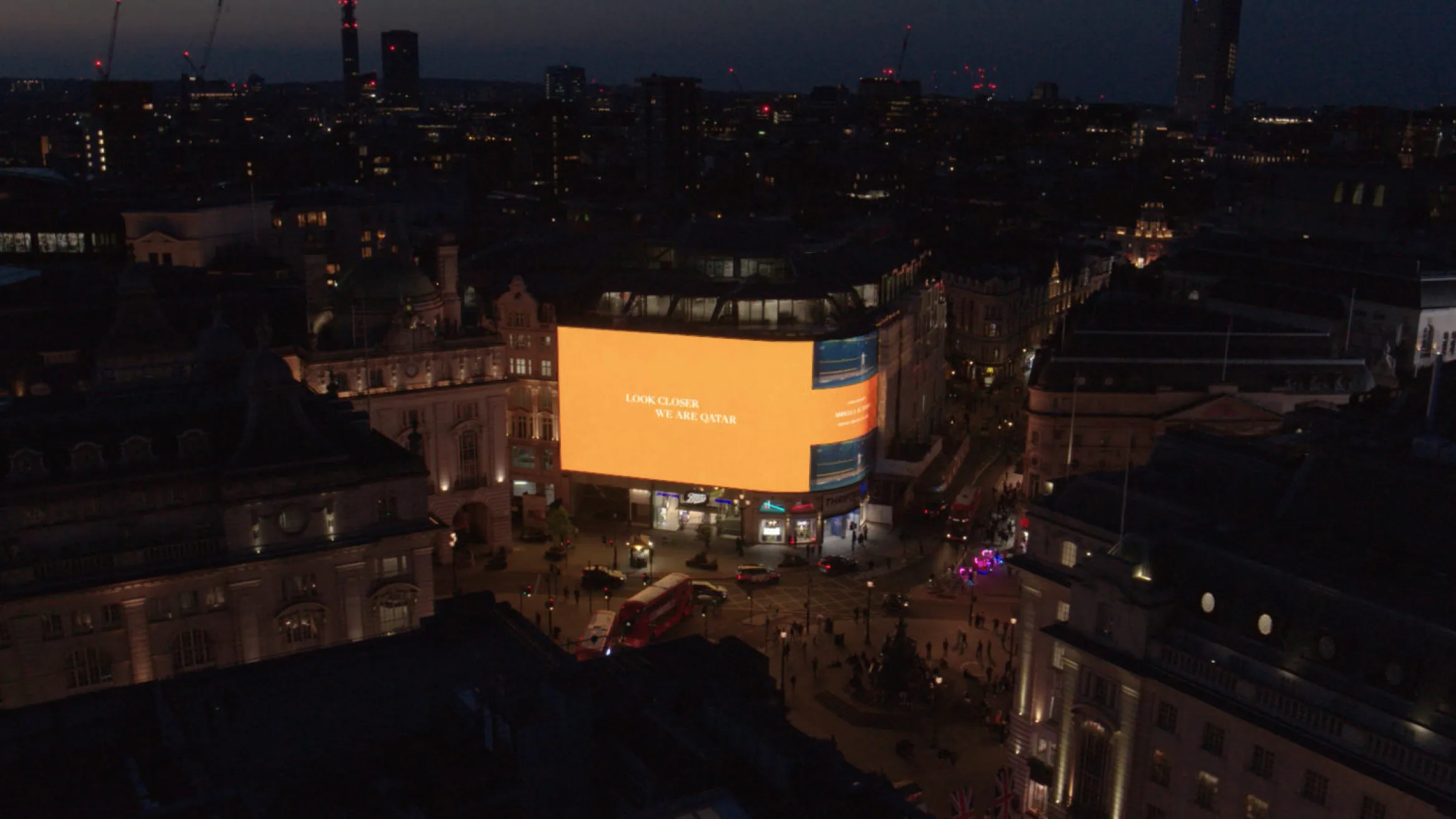 A nighttime view of Piccadilly Circus in London, featuring illuminated buildings and a large digital billboard glowing with an orange advertisement. The streets are bustling with vehicles and pedestrians, and city lights twinkle in the background.