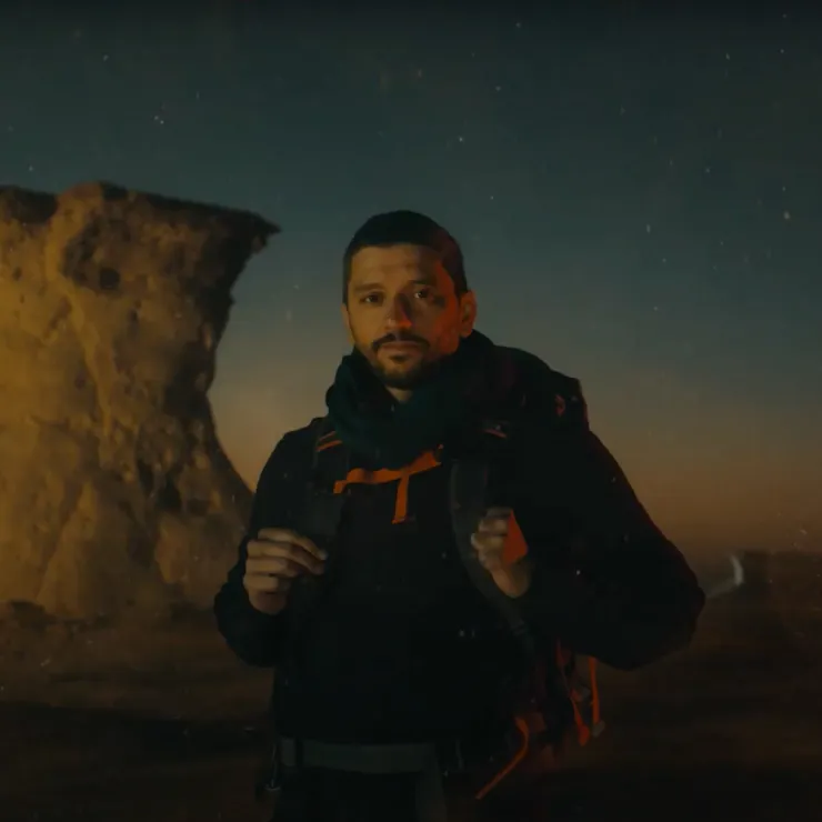 A person with a backpack stands outdoors at twilight with a rugged, cliff-like rock formation in the background. The person is wearing a dark jacket and scarf, looking directly at the camera. The sky is dimly lit, suggesting either dawn or dusk.