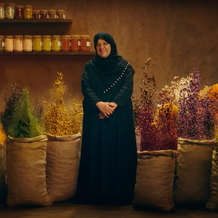 A woman in a black dress and headscarf stands smiling in front of a display of colorful dried flowers in large sacks. Shelves with jars of spices or preserves are seen in the background against a rustic wall.