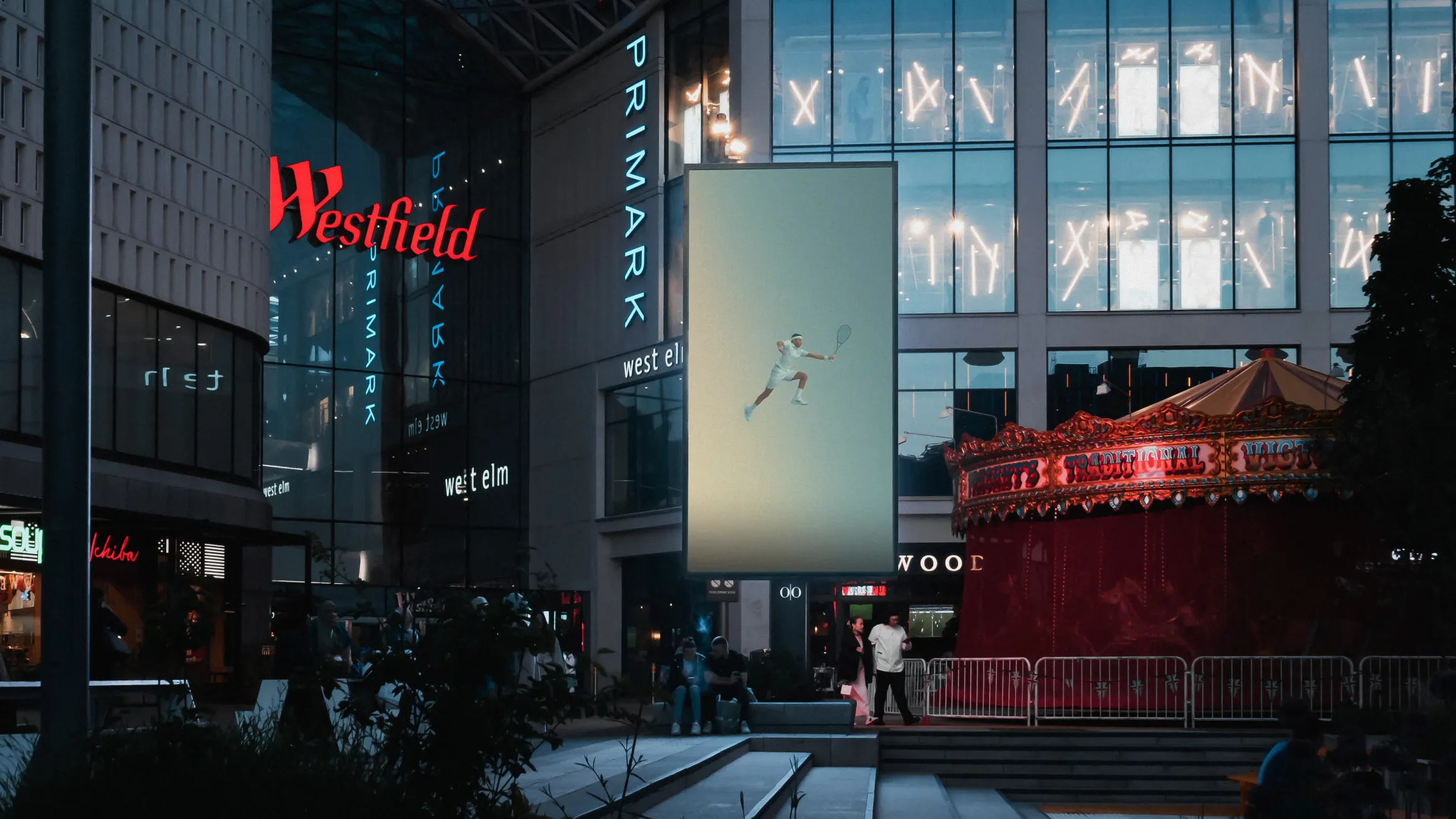 A bustling urban scene with people walking around a Westfield shopping center at dusk. The Primark store is prominently lit-up. A brightly illuminated video display screen features a basketball player dunking. There's a carousel with red canopy on the right.