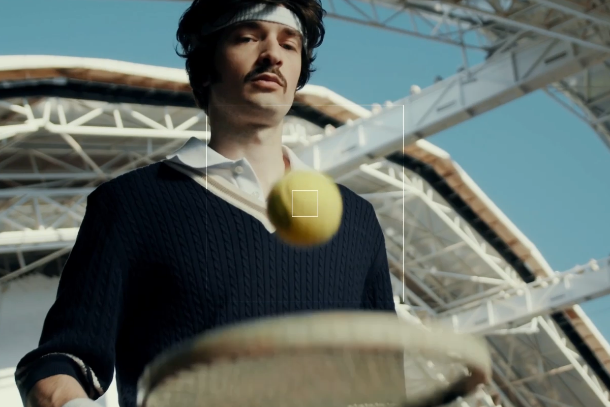 A person wearing a sweater and headband is poised to hit a tennis ball with a racket, looking as if they could star in any sizzle reel. The backdrop showcases a large, modern structure under the clear sky.