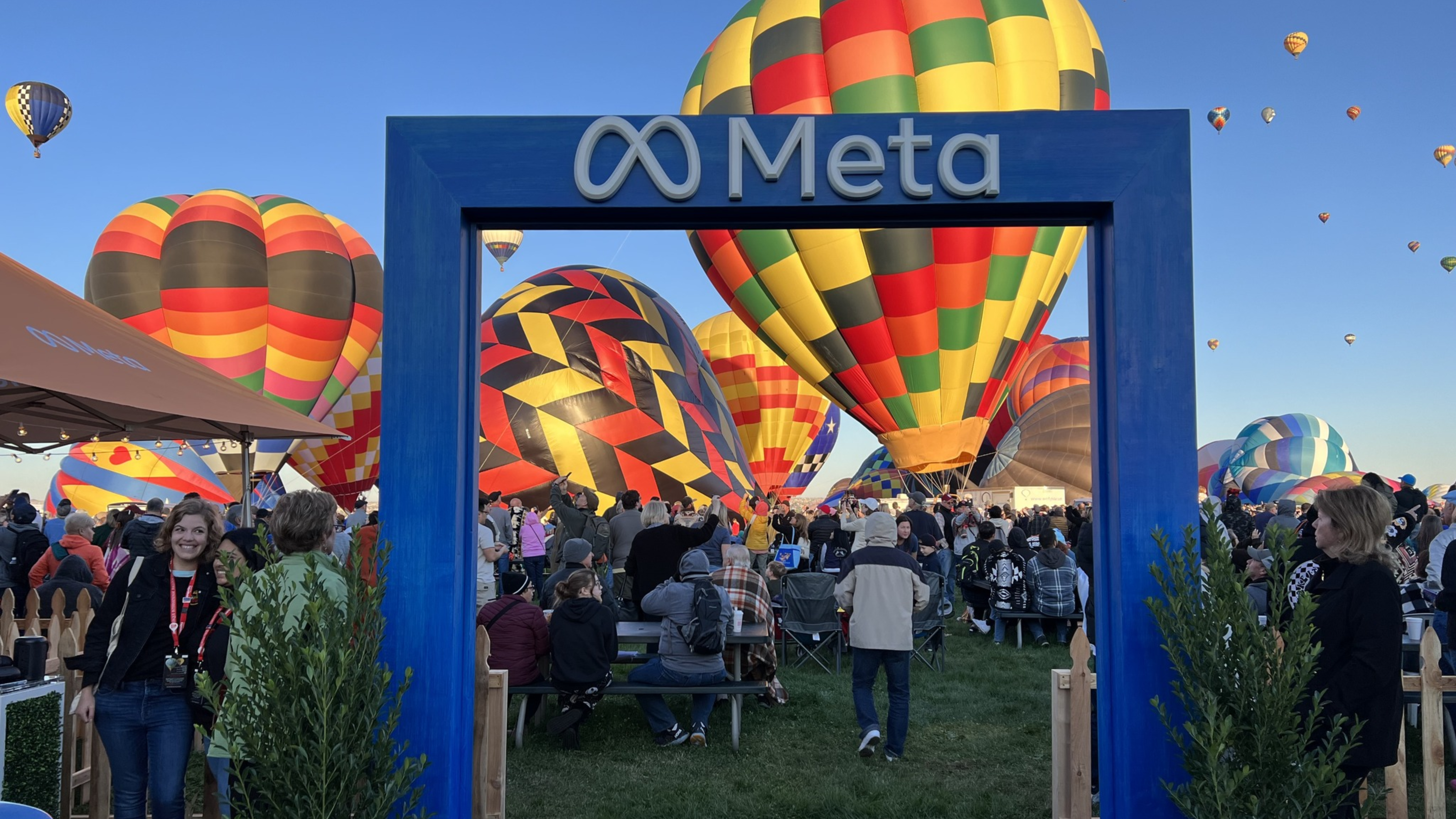 A crowd gathers at the hot air balloon festival as numerous colorful balloons fill the sky. In the foreground, a blue frame with "Meta" written on it stands like a portal to another world, drawing attention like a website redesign unveiling new horizons. People walk through and gather around in awe.