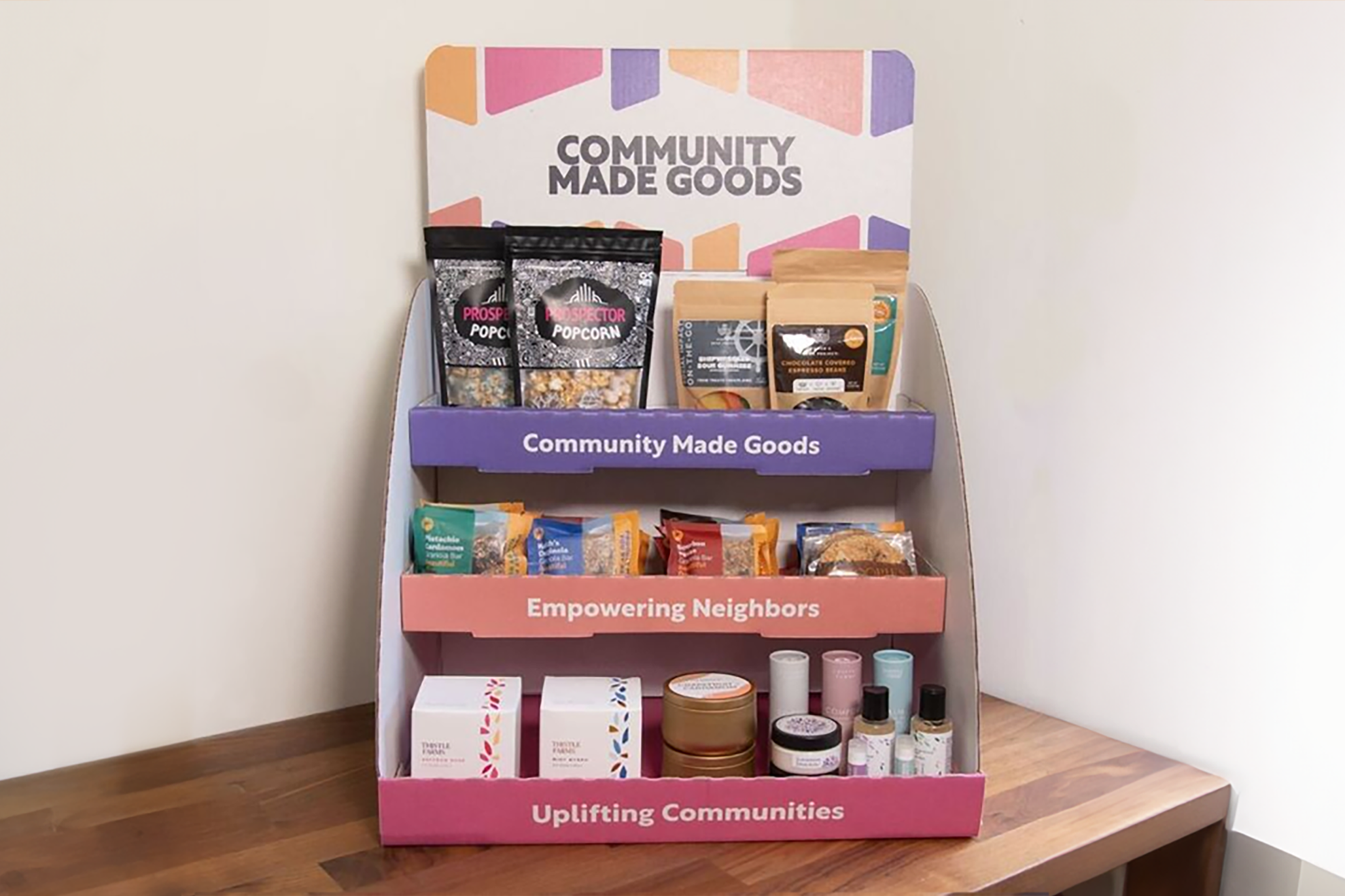 Colorful display stand with three shelves labeled "Community Made Goods," "Empowering Neighbors," and "Uplifting Communities," showcasing snacks, candles, soaps, and small products crafted by underrepresented groups. Sign on top reads "Community Made Goods.