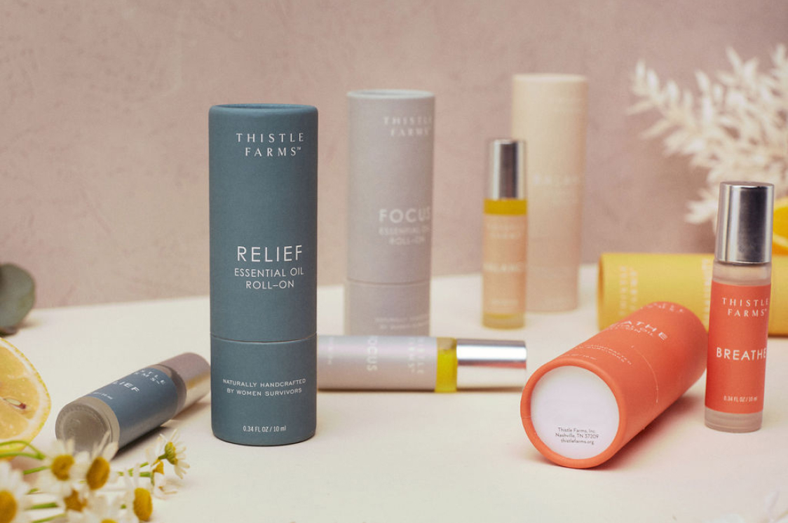 A collection of Thistle Farms essential oil products, including roll-ons labeled "Relief," "Focus," and "Breathe," elegantly displayed on a light surface. With flowers and a lemon slice nearby against a soft pink background, these products not only elevate wellness but also support social impact initiatives.