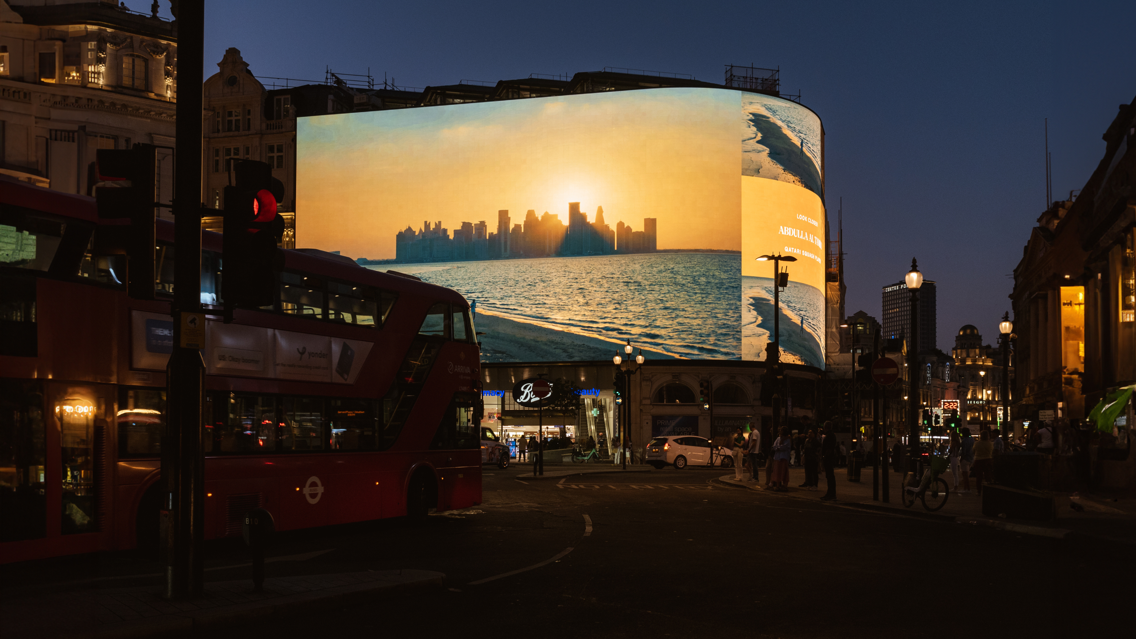 A busy city street at night showcases nation branding with a large, illuminated digital billboard displaying a sunset over the skyline. A red double-decker bus and several pedestrians are visible in the foreground, while streetlights and buildings glow vibrantly.