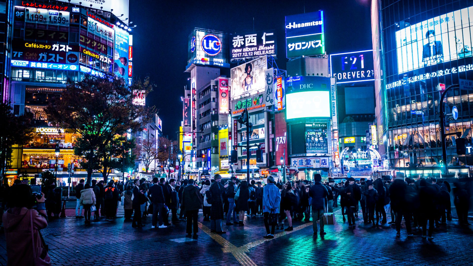 A bustling city scene at night featuring a crowded intersection illuminated by vibrant neon signs and billboards. Skyscrapers tower above, and the streets are filled with people crossing in all directions under the colorful lights.