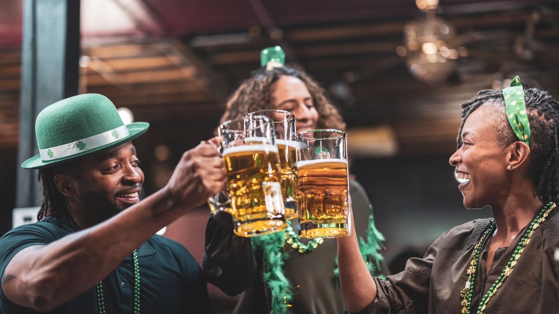 Three people celebrating and clinking mugs of beer, wearing festive green attire and accessories. They are smiling and appear to be enjoying a lively, cheerful atmosphere indoors.