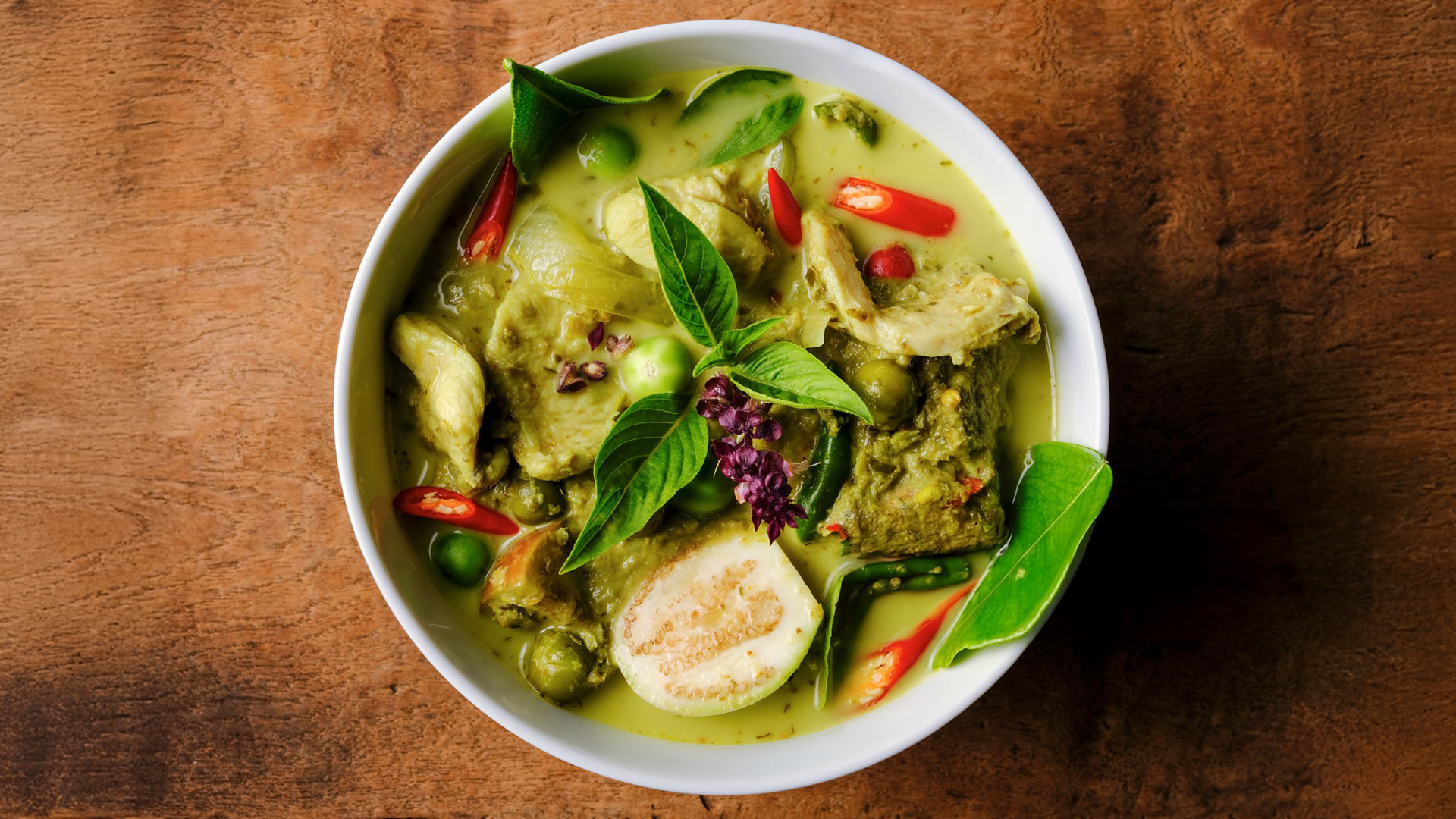 A bowl of vibrant green curry with vegetables and herbs, including eggplant slices, red chilies, and basil leaves, sits on a wooden table. The dish features a creamy, greenish-yellow curry sauce.