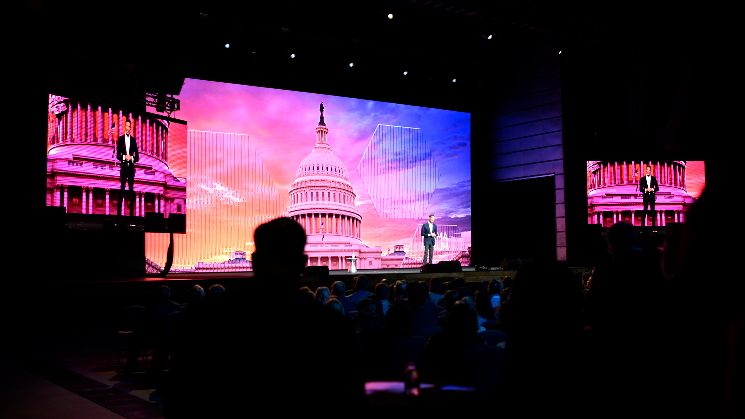 A speaker stands on stage in front of a large digital screen displaying an image of the U.S. Capitol with a vibrant sunset sky, discussing health communication strategies. The audience is silhouetted in the foreground, captivated by insights into effective public policy communications.