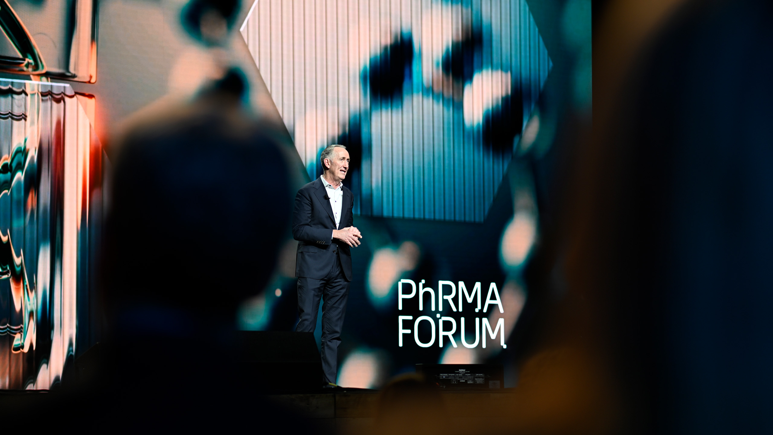 A person in a suit stands on stage speaking at the "PhRMA Forum," weaving elements of health communication into their presentation, against a backdrop of abstract blue and green visuals. Audience members engage eagerly in the foreground.