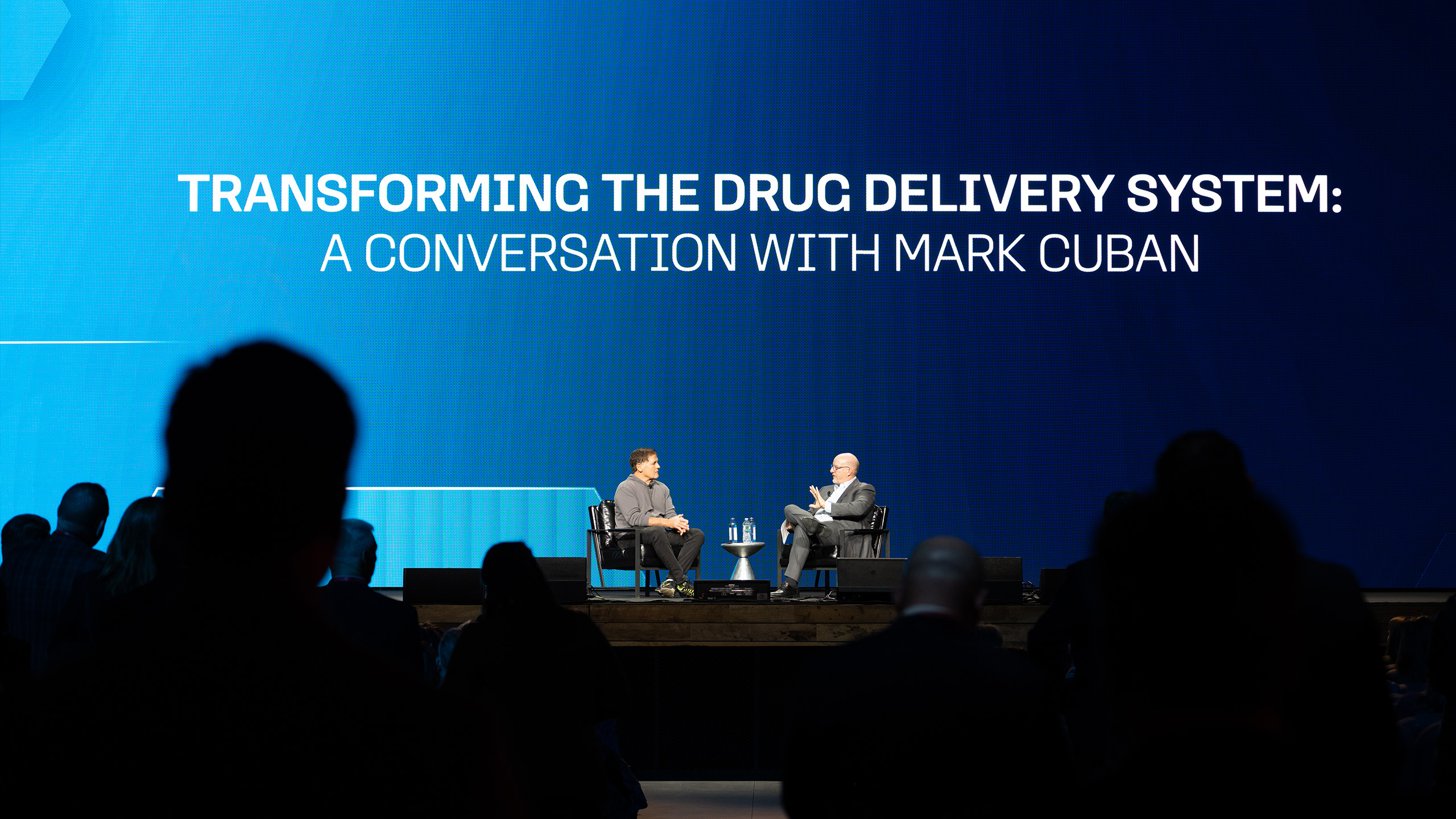 Two individuals are seated on stage, engaging in a compelling dialogue about transforming the drug delivery system. The large screen behind them reads "A Conversation with Mark Cuban." This insightful exchange highlights innovative health communication strategies, as an attentive audience looks on.