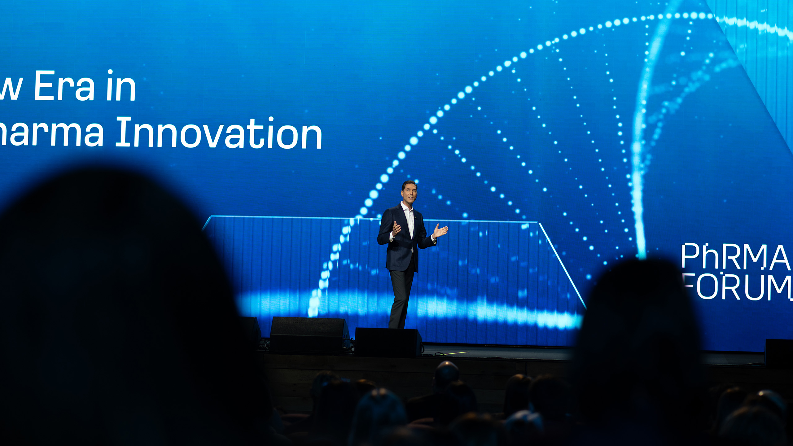 A speaker stands on stage at the Pharma Forum, with a large blue screen behind them displaying "New Era in Pharma Innovation" alongside a DNA helix graphic, underscoring a pivotal moment for health advocacy. The attentive audience is visible in the foreground, eager to engage in this dialogue.