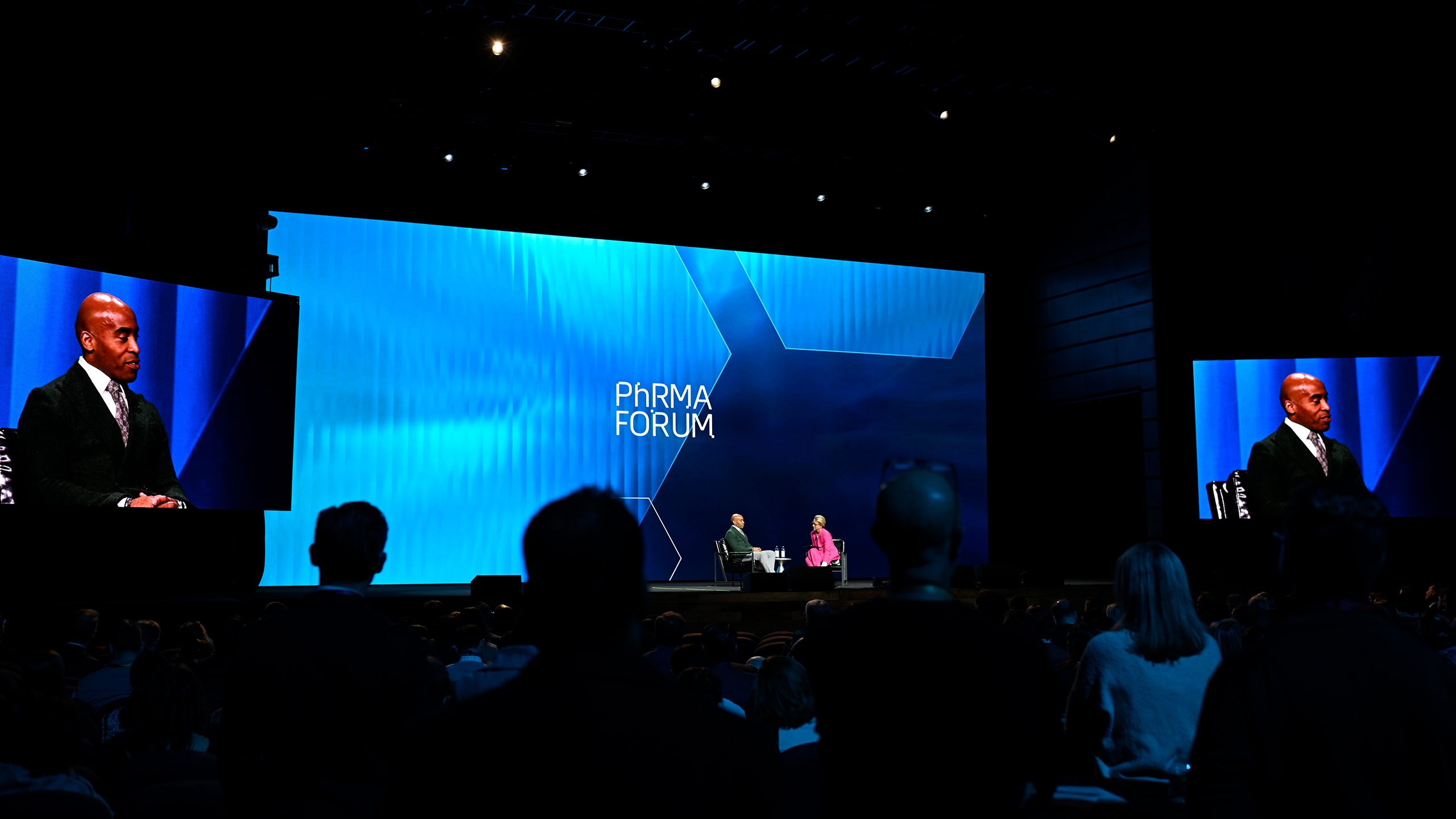 A large stage displays the words "Pharma Forum" with two people engaged in a dialogue on communication strategy. The audience is visible in the foreground, and large screens showcase speaker close-ups. The dimly lit setting creates an intimate, seminar-like atmosphere.