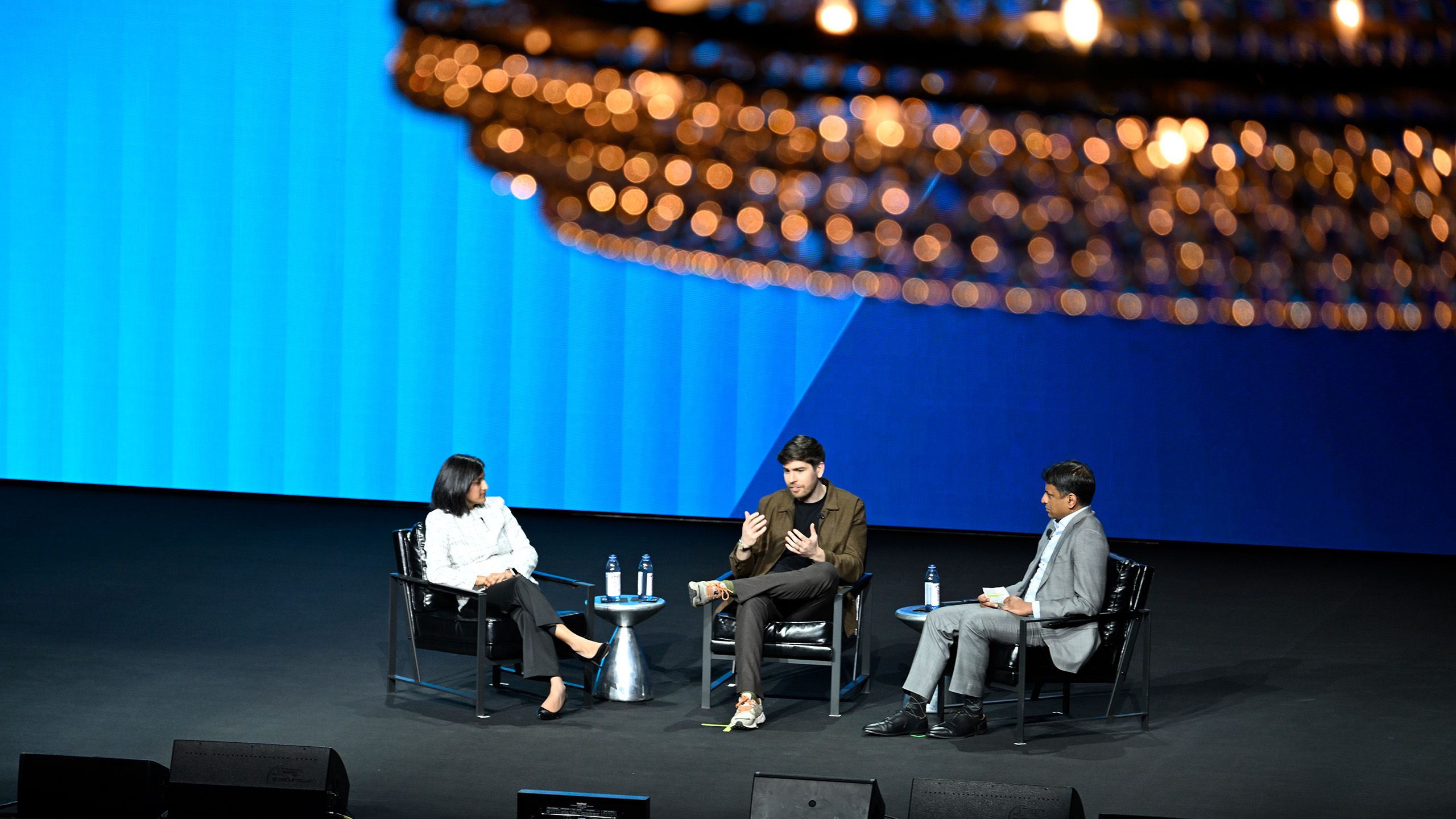 Three people sit on a stage with a blue backdrop, engaged in a panel discussion. Two men and a woman, focusing on health advocacy, are seated in armchairs with microphones. A large chandelier is partially visible above them.