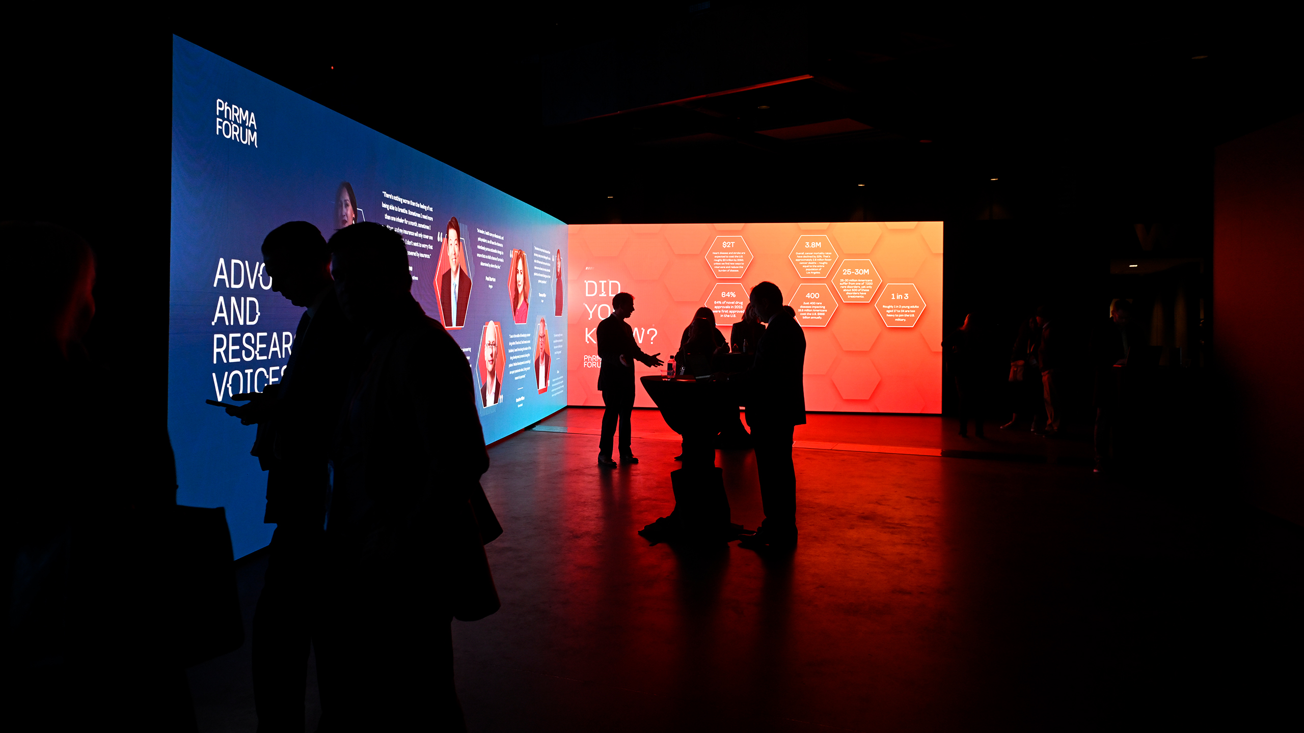 In a dimly lit room, silhouetted figures gather near illuminated digital screens, engaging in health communication. One screen glows blue with text, while another shines red with hexagonal patterns, as they converse or observe the dynamic displays.