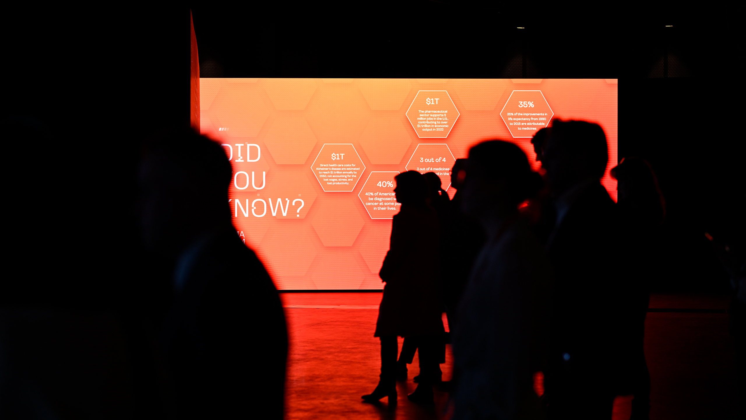 Silhouettes of people walking in front of a large screen displaying text and hexagonal shapes, reminiscent of an innovative health communication strategy. The background is illuminated in red and orange hues.