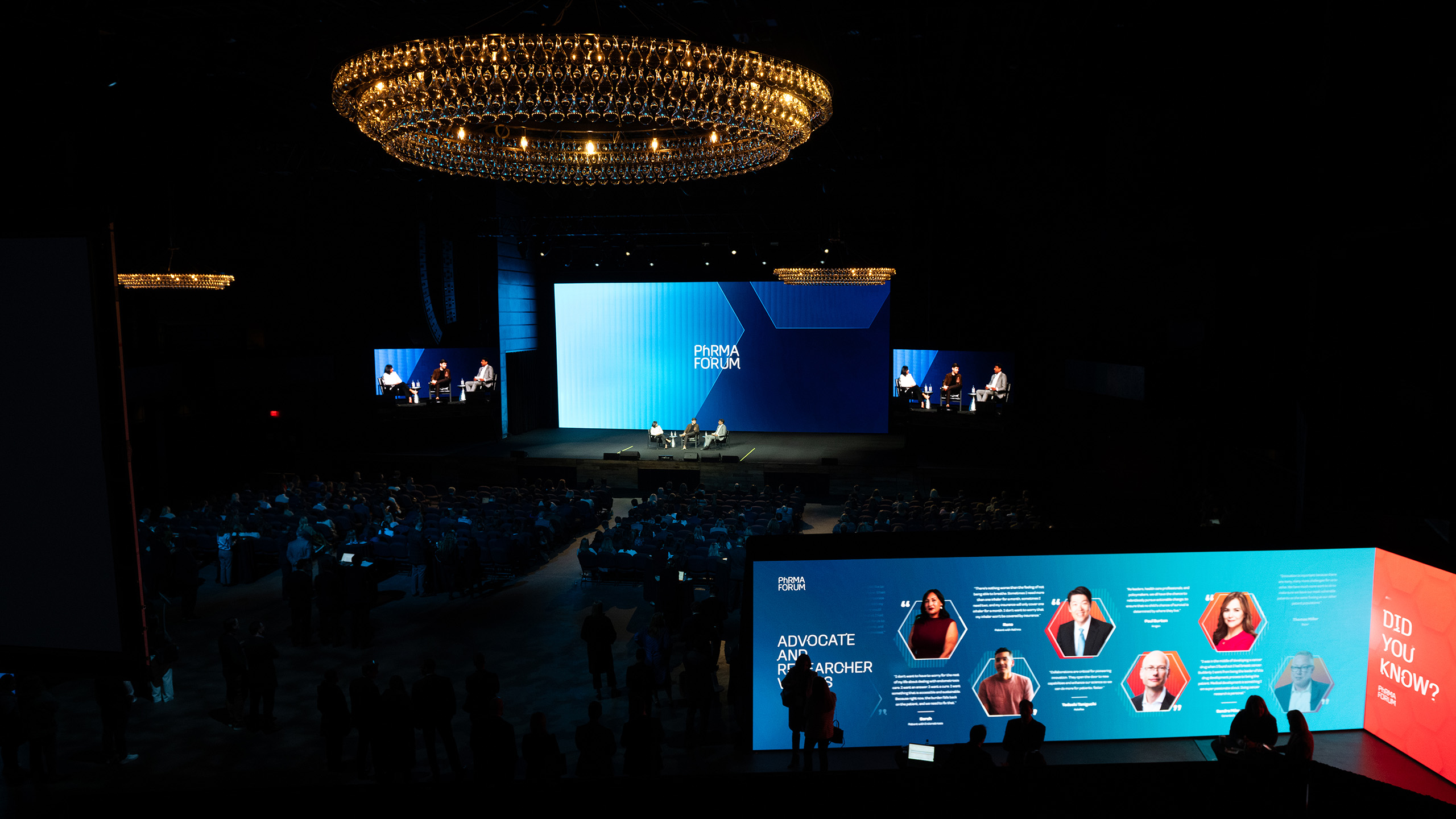 A large conference room with a brightly lit stage features two speakers engaged in a panel on health advocacy. Two screens display the event name while an LED wall showcases profiles. The audience listens intently, recognizing the impact of public policy communications in the dimly lit background.