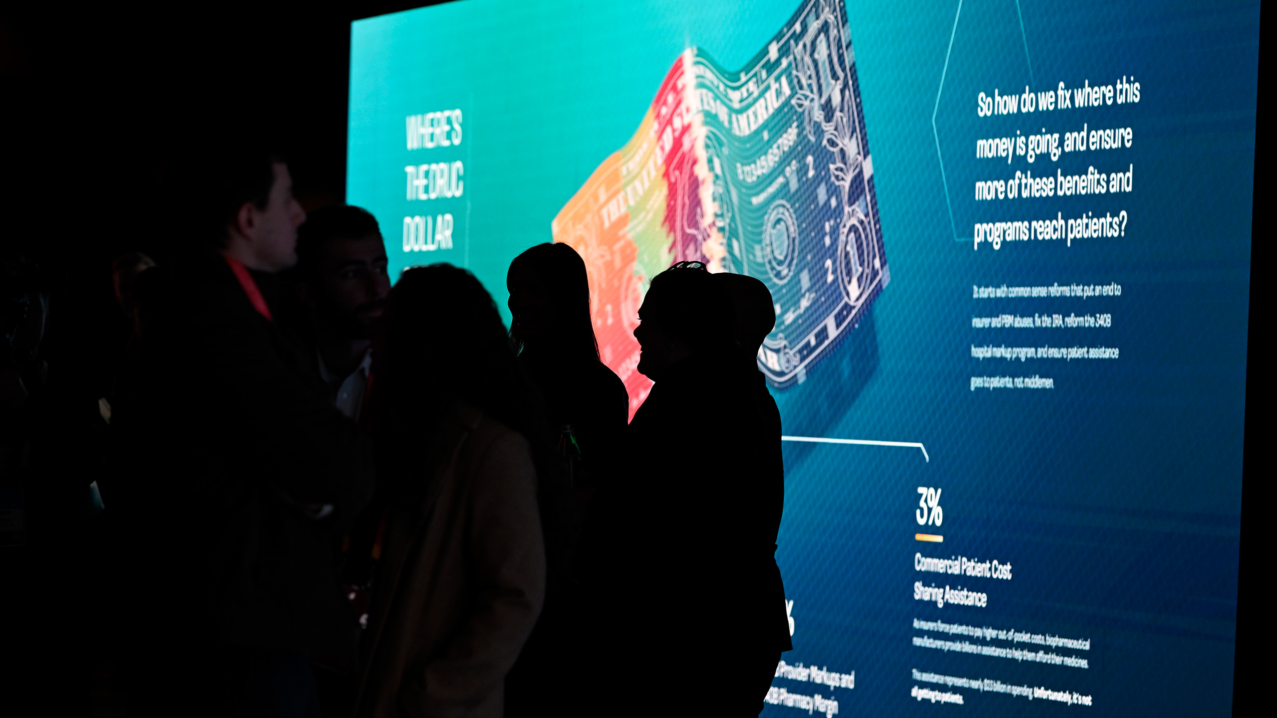 Silhouettes of people stand before a large, illuminated screen displaying vibrant images of currency and text on the impact of money, serving as the centerpiece in a dynamic digital campaign.