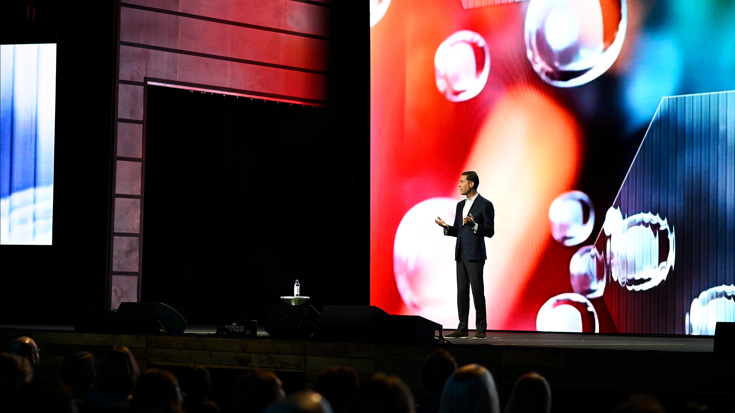 A person in a suit stands on stage delivering a presentation on health communication. Behind them, a large screen displays abstract colorful imagery with circular shapes. A bottle of water and a microphone stand are on the podium. An audience is visible in the foreground.