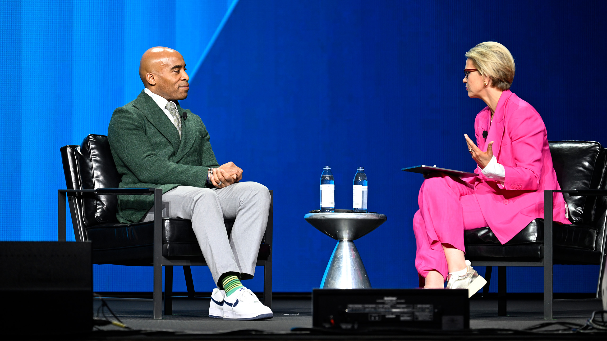 Two people are seated on stage engaging in a conversation about public policy communications. The person on the left wears a green blazer, gray pants, and white shoes. The person on the right dons a bright pink suit and has short blonde hair. A small table with water bottles sits between them.