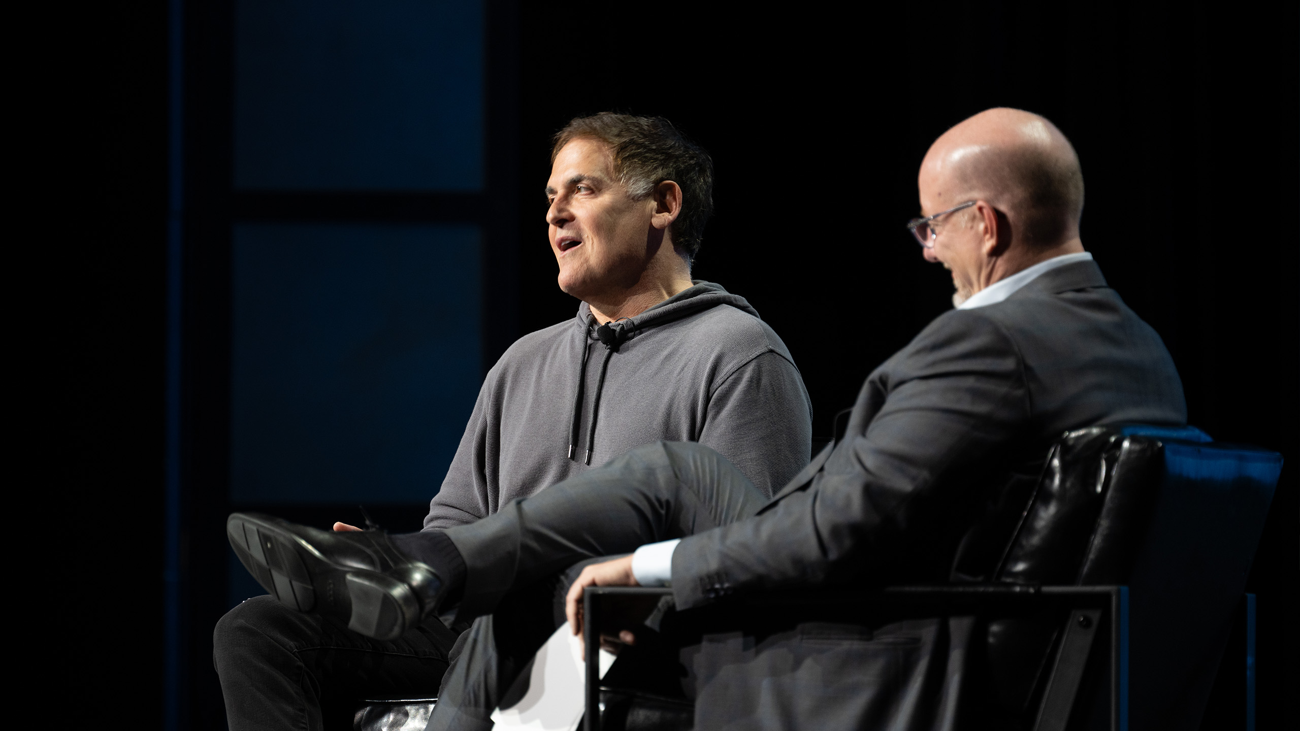 Two men are seated on a stage during a discussion. The man on the left, wearing a gray hoodie and discussing health advocacy, speaks, while the man on the right, in a suit, listens attentively. Both sit in black chairs against a dark background.