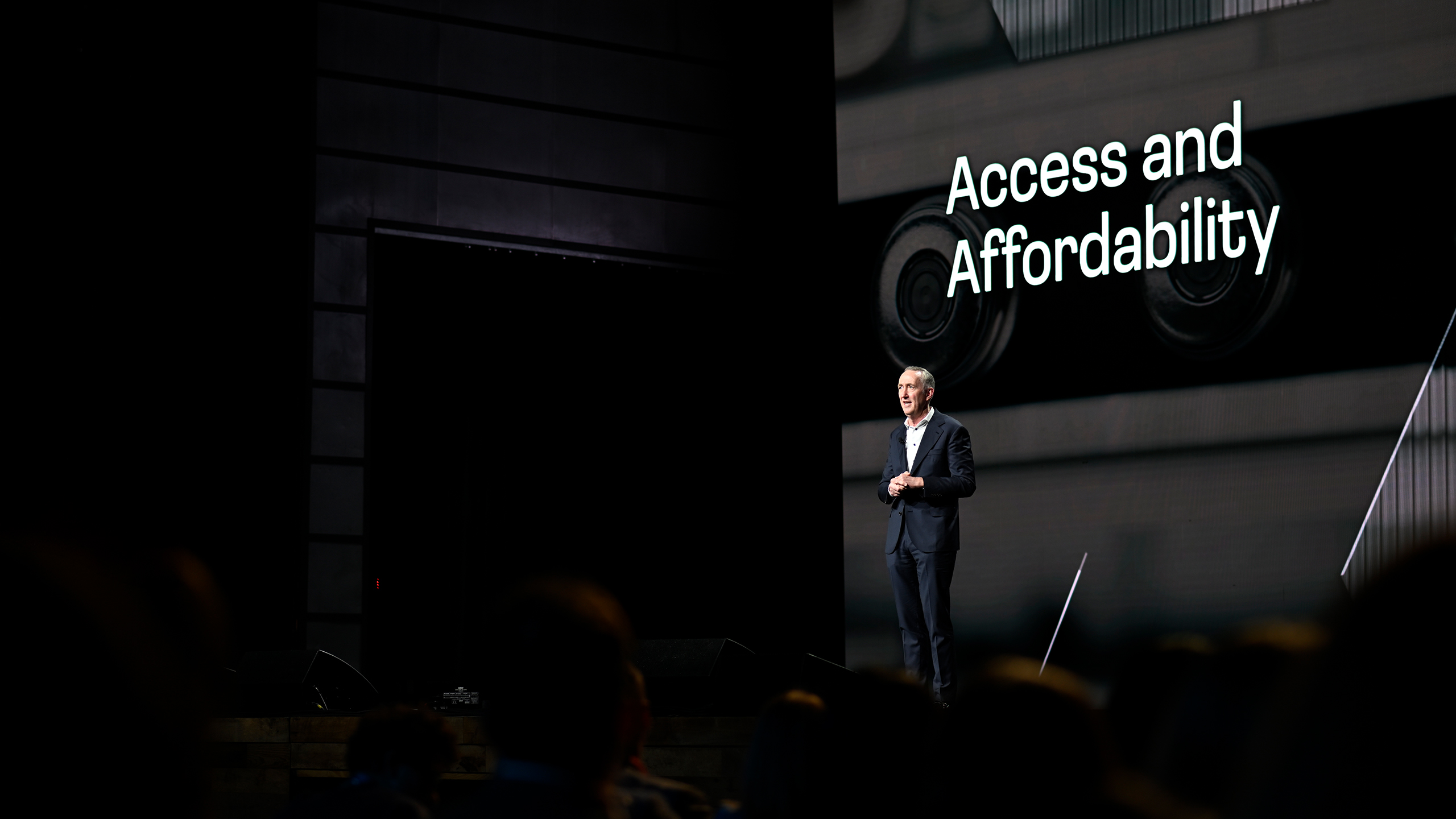 A man in a suit speaks on stage with "Access and Affordability" in large text behind him, emphasizing crucial health communication. Abstract black and white images set the backdrop, while attentive audience members are visible in the foreground.