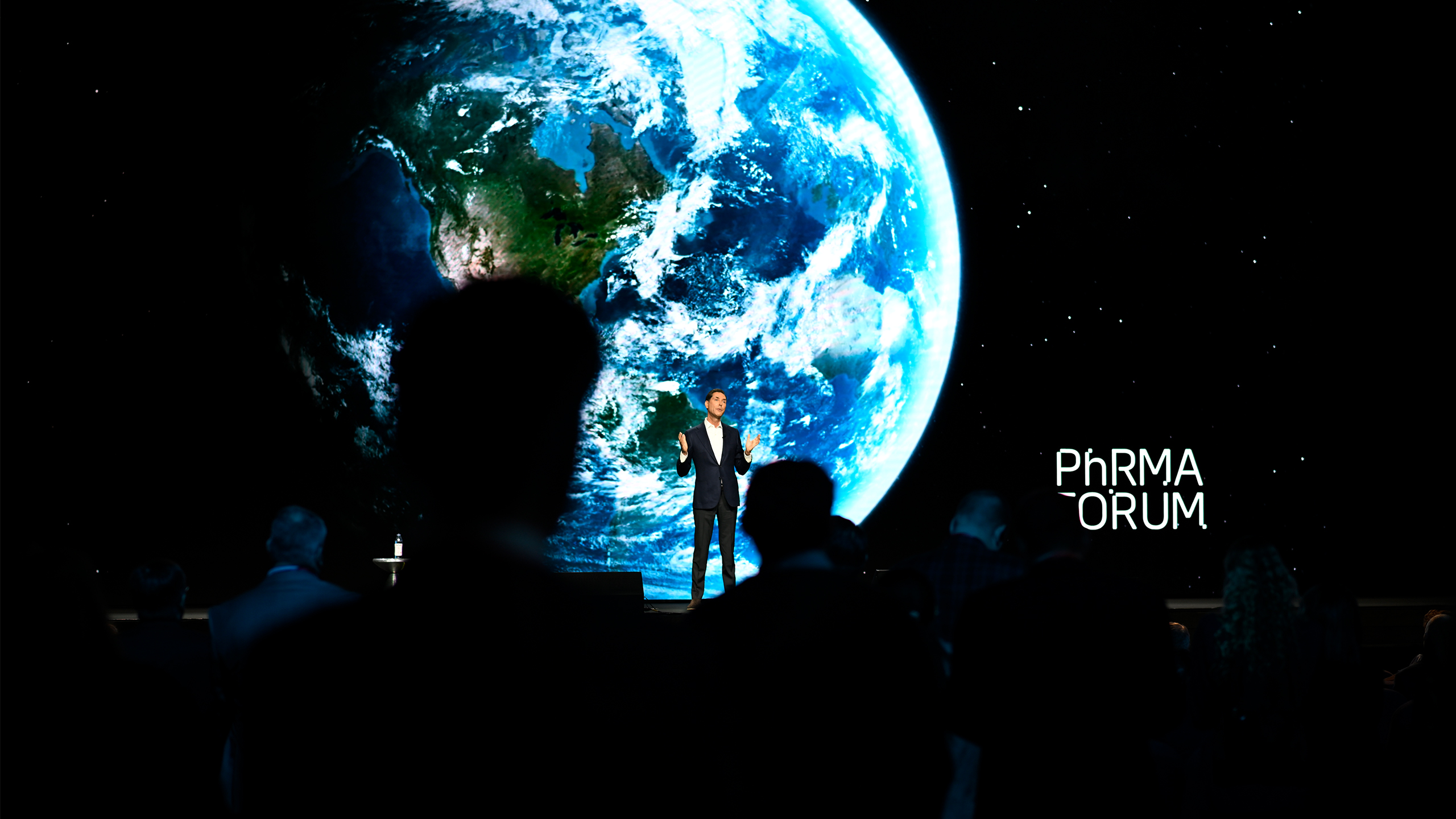 A person stands on stage at the "Pharma Forum," delivering insights on health advocacy, framed by a breathtaking projection of Earth from space. The audience listens intently, silhouetted against the vibrant blue and green colors of the planet.