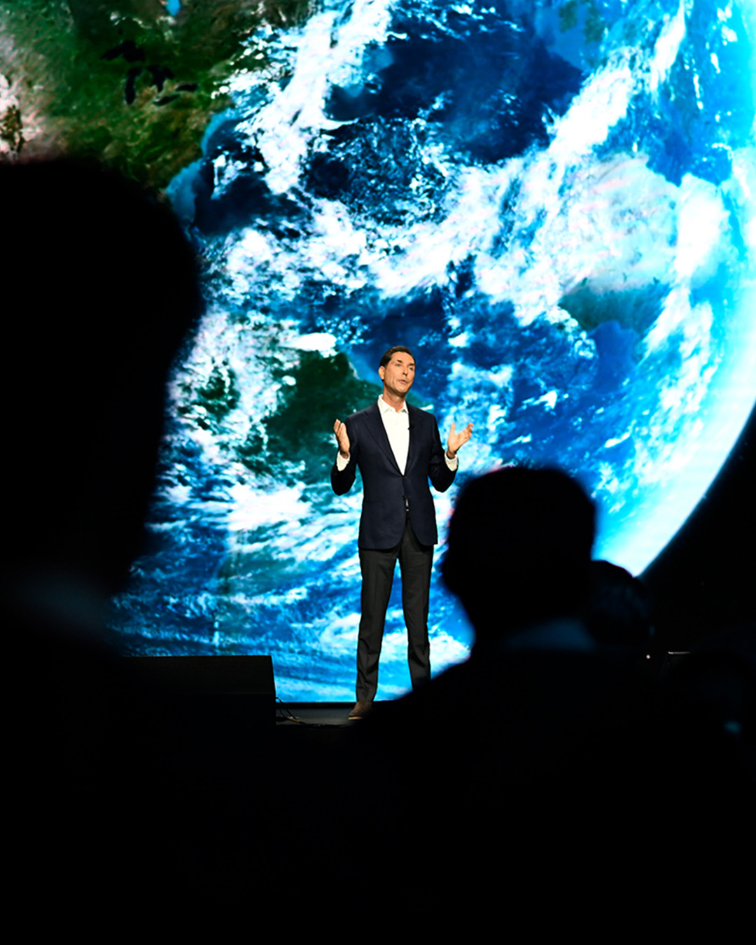 A person in a suit speaks on stage with a large image of Earth projected in the background, illustrating their innovative communication strategy. Audience members are silhouetted, enhancing the presentation's focus and impact.