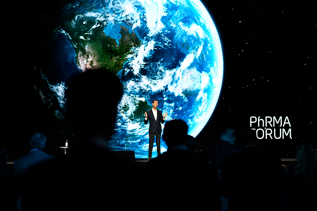 A person stands on stage with a large Earth image projected behind them, embodying Health Advocacy in action. The word "PhRMA ORUM" appears in the background, hinting at a conference focused on global health issues. Audience silhouettes are visible in the foreground, engaged and attentive.